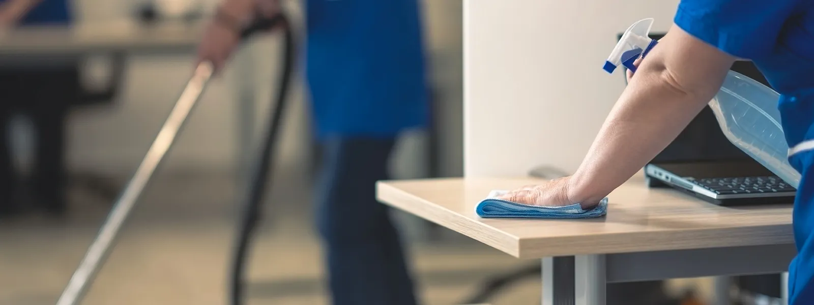 Lokalvårdare i blå arbetskläder torkar av ett skrivbord på kontor